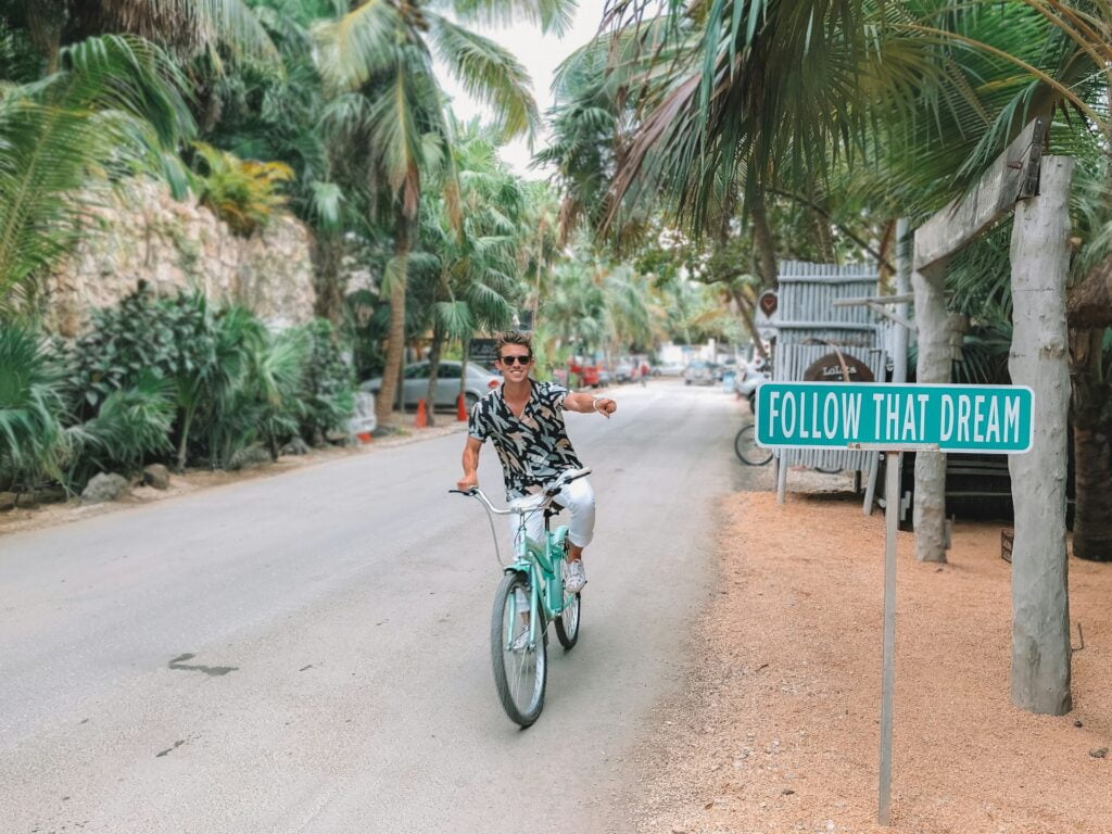 "Digital nomad riding a bike in Tulum, passing by an inspiring 'Follow Your Dreams' sign, symbolizing the essence of freedom and the pursuit of a lifestyle that blends work, adventure, and the serene beauty of Tulum's natural surroundings