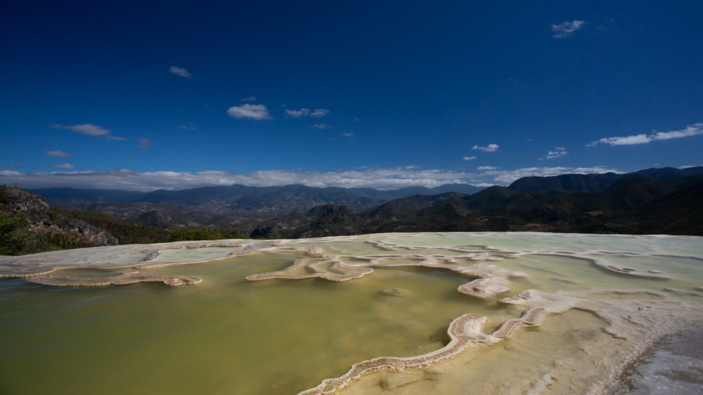 A serene freshwater pond in the foreground, reflecting the clear blue sky of Oaxaca, Mexico, with majestic mountains rising in the background, symbolizing the tranquil and natural beauty ideal for a rejuvenating retiremen